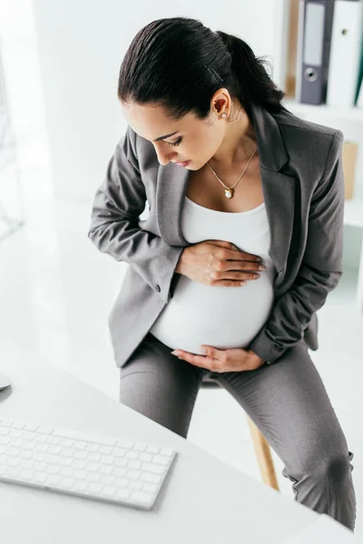 Visão de alto ângulo da mulher grávida sentada atrás da mesa e segurando a barriga — Fotografia de Stock