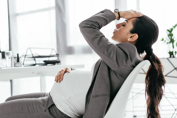 Tired pregnant woman sitting in office in chair near table and holding hear with hand — Stock Photo