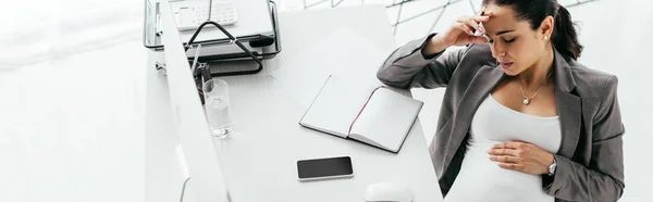 Panoramic shot of pregnant woman holding head with hand and sitting behind table with computer, notebook and document tray — Stock Photo