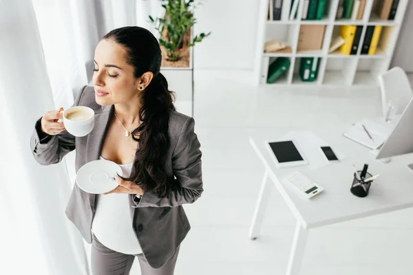 Высокий угол обзора беременной женщины, стоящей в офисе и пьющей кофе — стоковое фото
