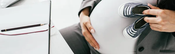 Панорамний знімок вагітної жінки, що тримає маленькі дитячі приманки, сидячи за столом з блокнотом — стокове фото