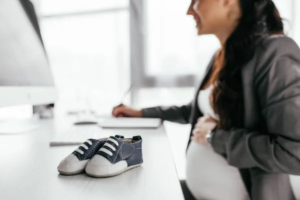 Foco seletivo de pequenas botas de bebê na mesa e mulher grávida trabalhando no computador — Fotografia de Stock