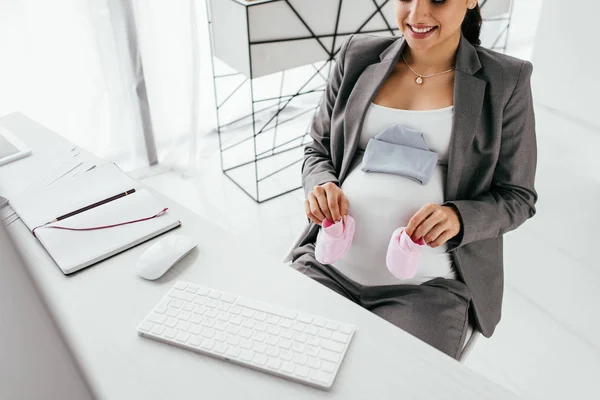 Mujer embarazada sentada detrás de la mesa con el teclado del ordenador y el ratón y la celebración de zapatillas de color rosa - foto de stock