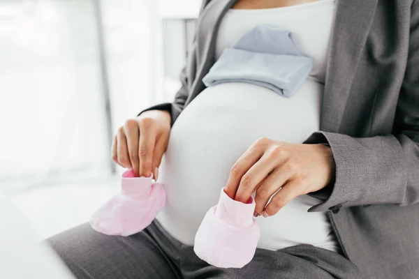 Cropped view of pregnant woman holding pink slippers and small hat on her belly — Stock Photo