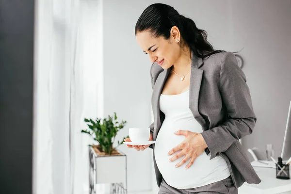 Schwangere steht mit Kaffeebecher im Büro und erträgt Schmerzen — Stockfoto