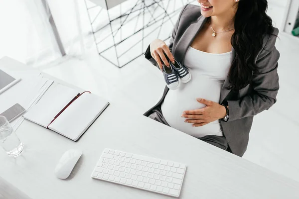 Ausgeschnittene Ansicht einer schwangeren Frau, die mit Computertastatur und -maus hinter einem Tisch sitzt und kleine Gummischuhe hält — Stockfoto