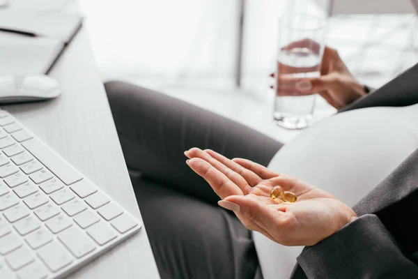 Vista recortada de la mujer embarazada sosteniendo píldoras de colores y vidrio con agua mientras está sentado detrás de la mesa - foto de stock