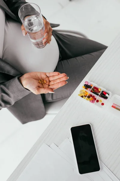 Vista superior de la mujer embarazada sosteniendo píldoras mientras está sentado detrás de la mesa con caja de pastillas - foto de stock