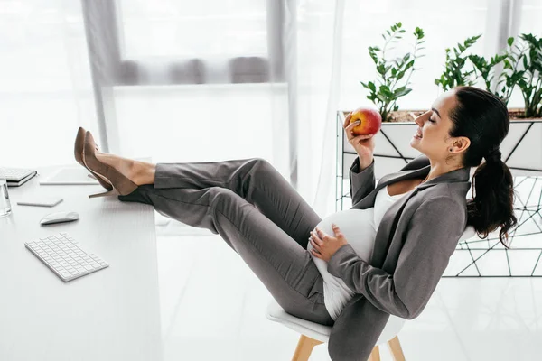 Вид збоку вагітної жінки тримає яблуко, сидячи в офісі — стокове фото
