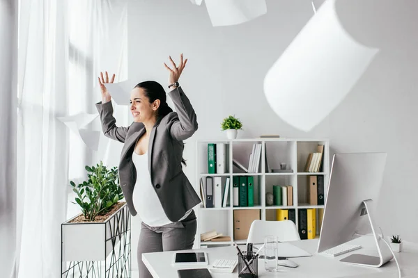 Раздраженная беременная женщина бросает бумаги в воздух, стоя у стола в офисе — стоковое фото