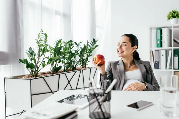Счастливая беременная женщина ест яблоко, сидя в офисе возле стола — стоковое фото