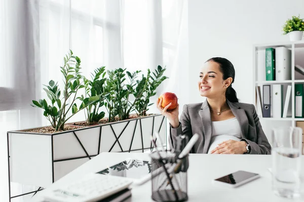 Feliz embarazada comer manzana mientras está sentado en la oficina cerca de la mesa - foto de stock