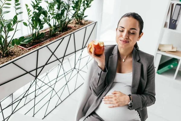 Femme enceinte mangeant de la pomme alors qu'elle est assise au bureau — Photo de stock