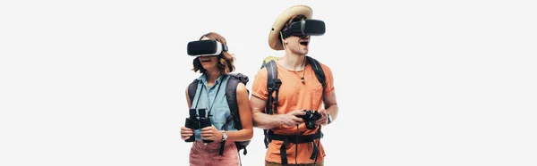 Tiro panorâmico de dois jovens turistas com binóculos e câmera digital usando fones de ouvido de realidade virtual isolados em cinza — Fotografia de Stock