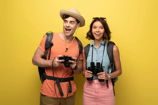 Two smiling tourists holding digital camera and binoculars on yellow background — Stock Photo