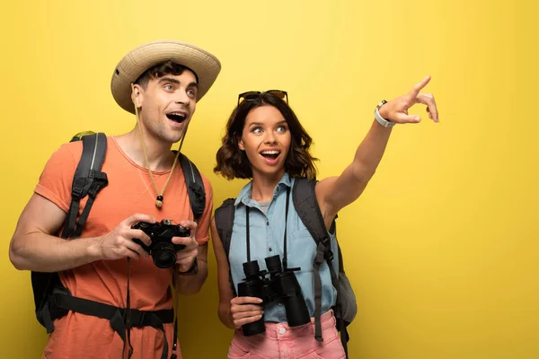 Fröhliche junge Frau, die wegschaut und mit dem Finger zeigt, während sie neben einem überraschten Mann auf gelbem Hintergrund steht — Stockfoto