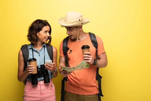 Sonriente mujer joven sosteniendo teléfono inteligente mientras mira al hombre con mapa geográfico sobre fondo amarillo - foto de stock