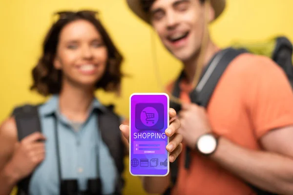 Selektiver Fokus einer fröhlichen Frau, die Smartphone mit Shopping-App zeigt, während sie neben einem lächelnden Mann auf gelbem Hintergrund steht — Stockfoto