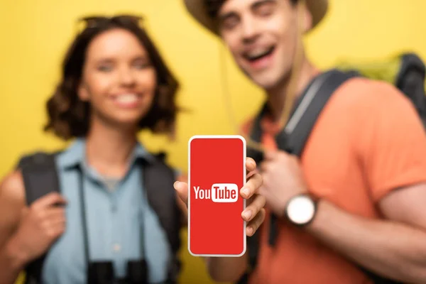 KYIV, UCRANIA - 3 de junio de 2019: enfoque selectivo de la mujer sonriente que muestra el teléfono inteligente con la aplicación Youtube mientras está cerca de un hombre alegre sobre un fondo amarillo . — Stock Photo