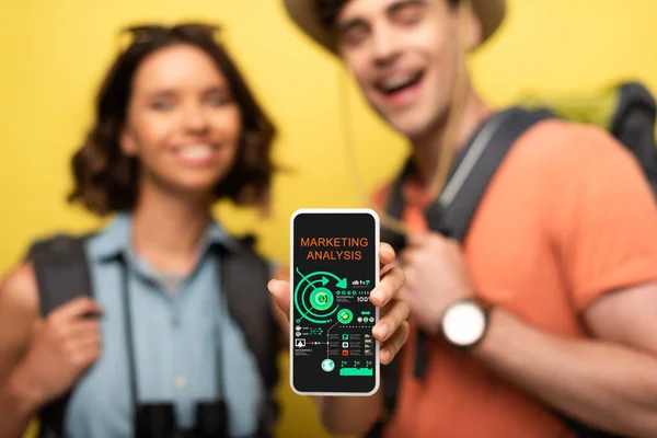 Селективное внимание веселой женщины, показывающей смартфон с приложением маркетингового анализа, стоя рядом с улыбающимся мужчиной на желтом фоне — стоковое фото
