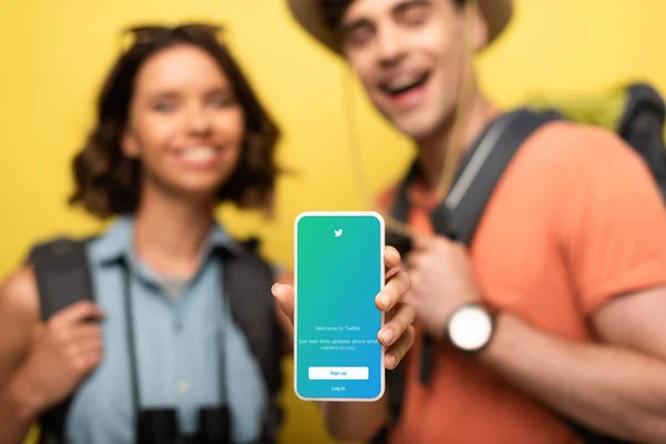 KYIV, UCRAINA - 3 GIUGNO 2019: Focus selettivo della donna sorridente che mostra lo smartphone con l'app Twitter mentre si trova vicino all'uomo allegro su sfondo giallo . — Foto stock