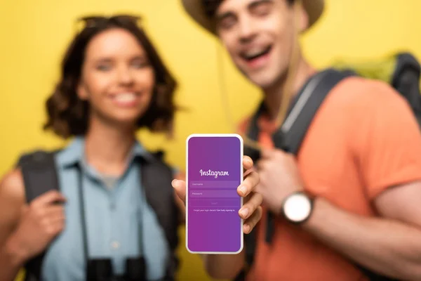 КИЕВ, УКРАИНА - 3 ИЮНЯ 2019: Выборочный фокус улыбающейся женщины, показывающей смартфон с приложением Instagram, стоя рядом с веселым мужчиной на желтом фоне . — стоковое фото