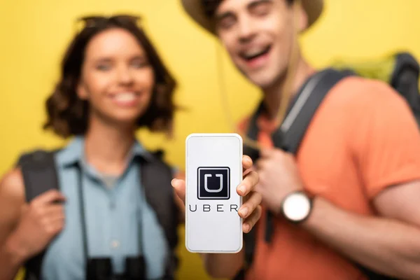 KYIV, UCRAINA - 3 GIUGNO 2019: Focus selettivo della donna allegra che tiene lo smartphone con l'app UBER mentre è in piedi vicino all'uomo sorridente su sfondo giallo . — Foto stock