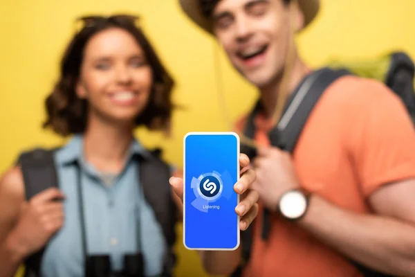 KYIV, UKRAINE - JUNE 3, 2019: Selective focus of cheerful woman holding smartphone with Shazam app while standing near smiling man on yellow background. — Stock Photo