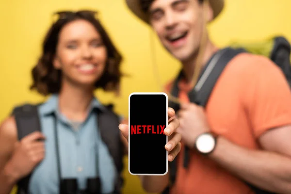 KYIV, UCRANIA - 3 de junio de 2019: enfoque selectivo de la mujer sonriente que muestra el teléfono inteligente con la aplicación Netflix mientras está cerca de un hombre alegre sobre un fondo amarillo . - foto de stock