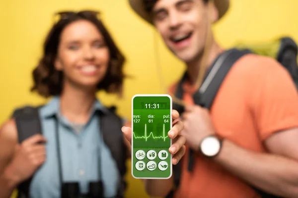 Selektiver Fokus einer fröhlichen Frau, die Smartphone mit Herzfrequenz-App zeigt, während sie neben einem lächelnden Mann auf gelbem Hintergrund steht — Stockfoto