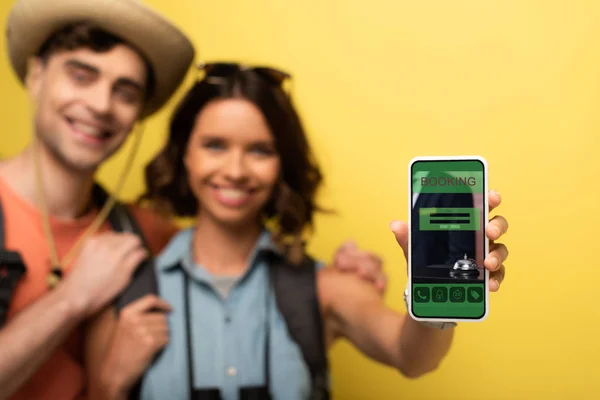 Selektiver Fokus eines lächelnden Mädchens, das neben einem fröhlichen Freund steht und Smartphone mit Buchungs-App auf gelbem Hintergrund zeigt — Stockfoto
