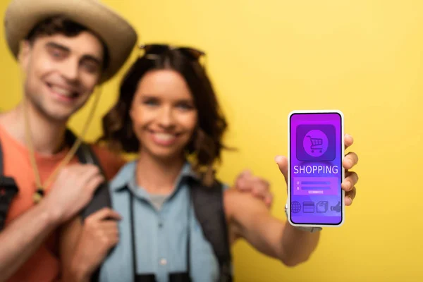 Foco seletivo de alegre jovem mulher de pé perto do namorado sorridente e mostrando smartphone com aplicativo de compras no fundo amarelo — Fotografia de Stock