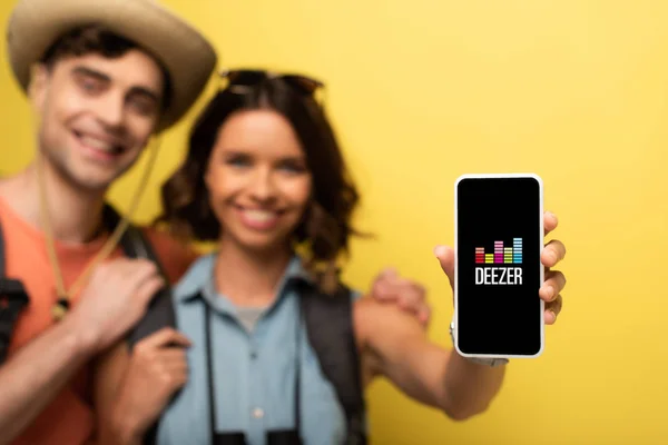 КИЕВ, УКРАИНА - 3 ИЮНЯ 2019 года: выборочное внимание веселой молодой женщины, стоящей рядом с улыбающимся парнем и показывающей смартфон с приложением Deezer на желтом фоне . — стоковое фото