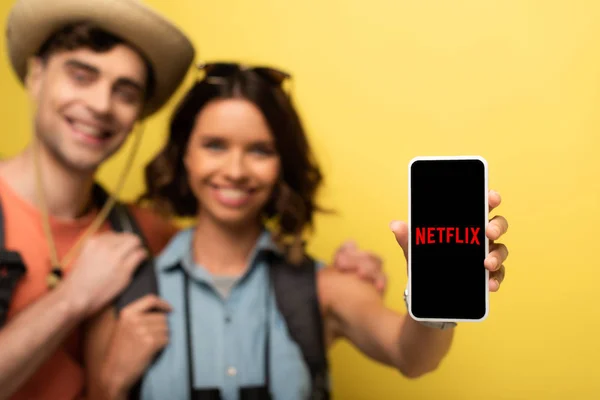 KYIV, UKRAINE - JUNHO 3, 2019: Foco seletivo de uma jovem alegre que está perto do namorado sorridente e mostra o smartphone com o aplicativo Netflix no fundo amarelo . — Fotografia de Stock