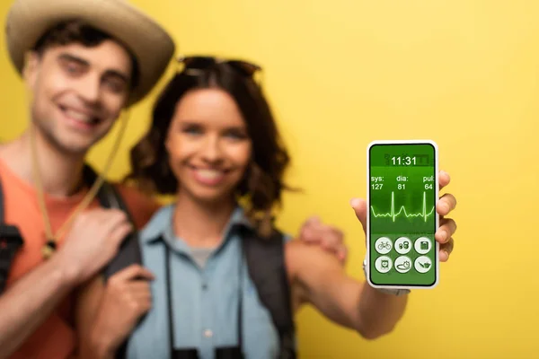 Selektiver Fokus einer fröhlichen jungen Frau, die neben ihrem lächelnden Freund steht und Smartphone mit Herzfrequenz-App auf gelbem Hintergrund zeigt — Stockfoto
