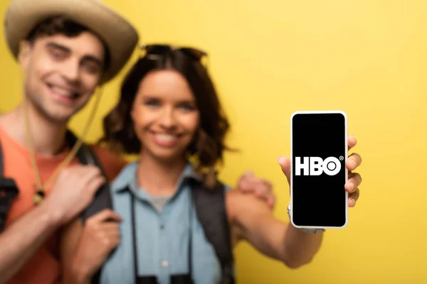 KYIV, UCRANIA - 3 DE JUNIO DE 2019: Enfoque selectivo de una joven alegre parada cerca de un novio sonriente y mostrando el teléfono inteligente con la aplicación HBO sobre fondo amarillo . - foto de stock