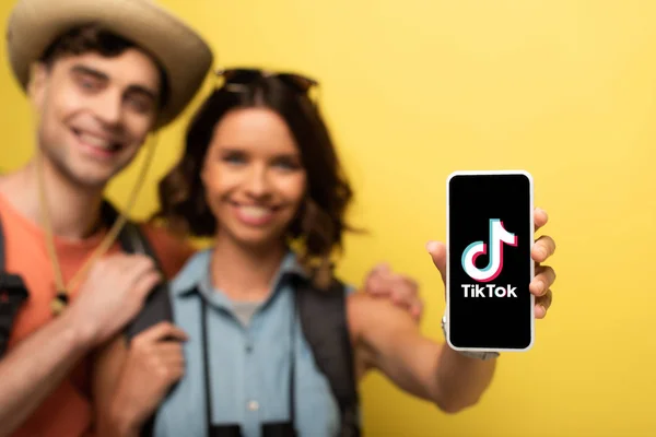 KYIV, UCRAINA - GIUGNO 3, 2019: Focus selettivo di giovane donna allegra in piedi vicino al fidanzato sorridente e mostrando lo smartphone con l'app Tik Tok su sfondo giallo . — Foto stock