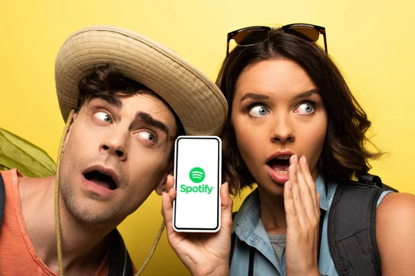 KYIV, UCRANIA - 3 de junio de 2019: Mujer joven sorprendida mostrando el teléfono inteligente con la aplicación Spotify mientras está cerca de un hombre sorprendido sobre un fondo amarillo . - foto de stock