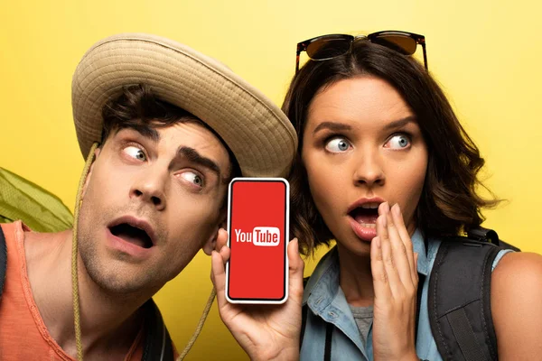 KYIV, UKRAINE - JUNE 3, 2019: Surprised young woman showing smartphone with Youtube app while standing near shocked man on yellow background. — Stock Photo