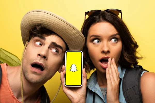 KYIV, UKRAINE - JUNHO 3, 2019: Jovem chocada mostrando smartphone com aplicativo Snapchat enquanto estava perto de homem surpreso no fundo amarelo . — Fotografia de Stock