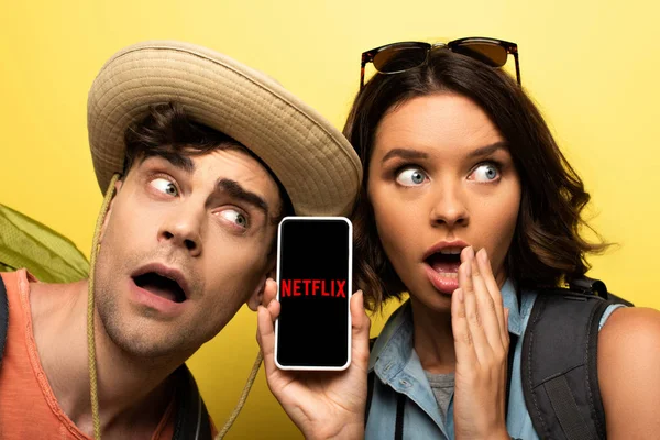 KYIV, UCRANIA - 3 de junio de 2019: Mujer joven sorprendida mostrando el teléfono inteligente con la aplicación Netflix mientras está cerca del hombre sorprendido sobre un fondo amarillo . - foto de stock