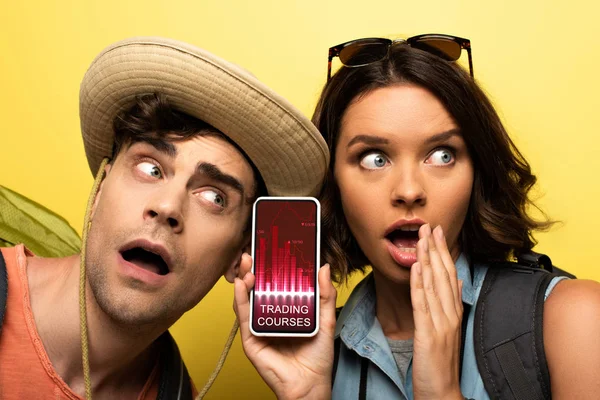 Chocado jovem mulher mostrando smartphone com cursos de negociação app enquanto estava perto homem surpreso no fundo amarelo — Fotografia de Stock