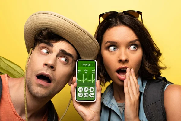 Schockierte junge Frau zeigt Smartphone mit Trading-Herzfrequenz-App, während sie neben einem überraschten Mann auf gelbem Hintergrund steht — Stockfoto