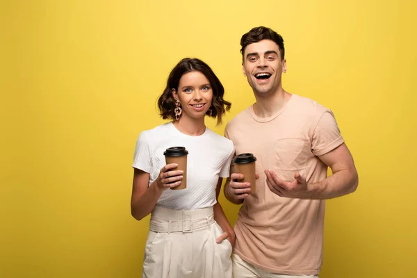 Homme et femme joyeux tenant café pour aller tout en souriant un appareil photo sur fond jaune — Photo de stock