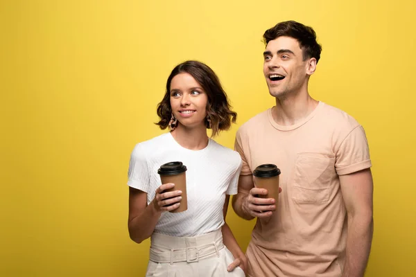Веселый мужчина и женщина держа одноразовые чашки, глядя в сторону на желтом фоне — стоковое фото