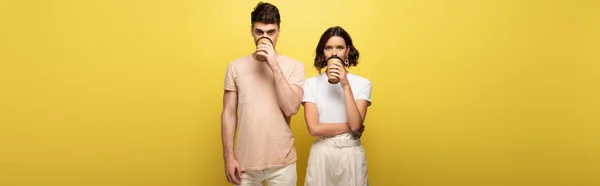 Panoramic shot of young man and woman drinking coffee to go while looking at camera on yellow background — Stock Photo