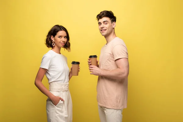 Smiling man and woman looking at camera while holding coffee to go on yellow background — Stock Photo