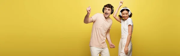 Panoramic shot of excited man and woman dancing while listening music in headphones on yellow background — Stock Photo