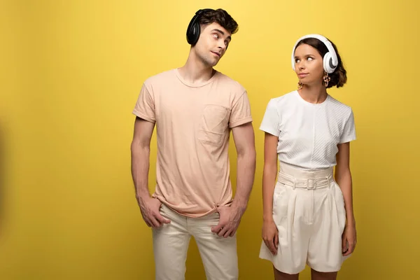 Joven hombre y mujer escuchando música en auriculares mientras se miran el uno al otro sobre un fondo amarillo - foto de stock