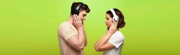 Panoramic shot of positive man and woman in headphones listening music with closed eyes on green background — Stock Photo